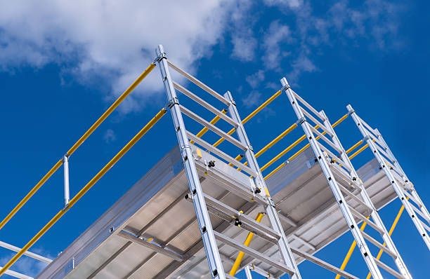 Aluminum construction scaffolding with yellow safety rails against a bright blue sky with a few clouds.